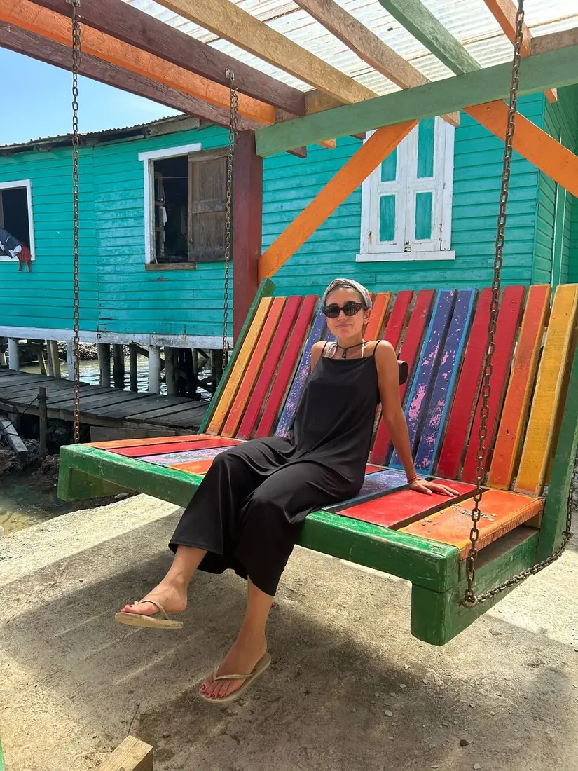 Viajera sentada en un columpio de madera de colores junto a una casa turquesa sobre el agua