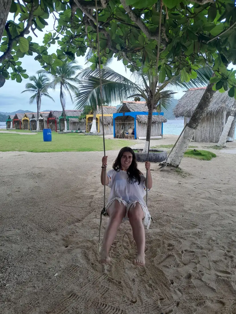 Viajera en un columpio bajo un árbol frente a cabañas de colores junto al mar