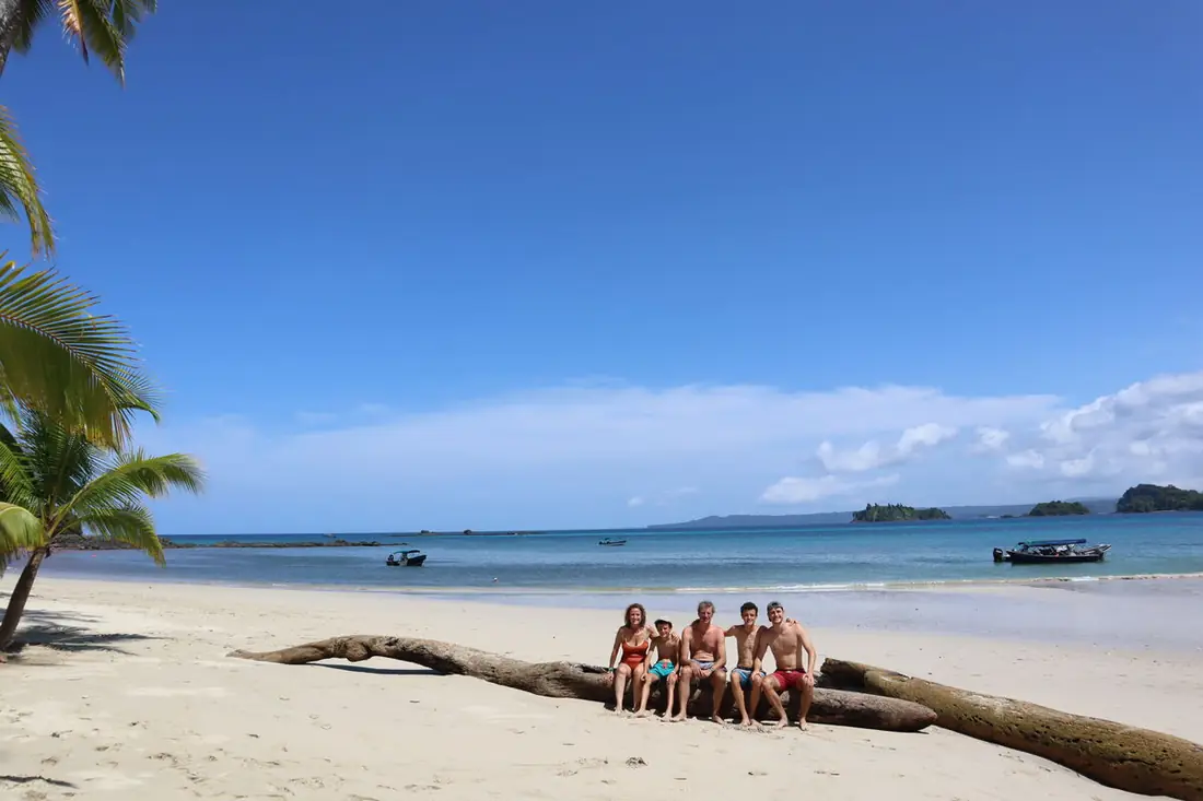 Familia sentada en un tronco en una playa de arena blanca y agua turquesa